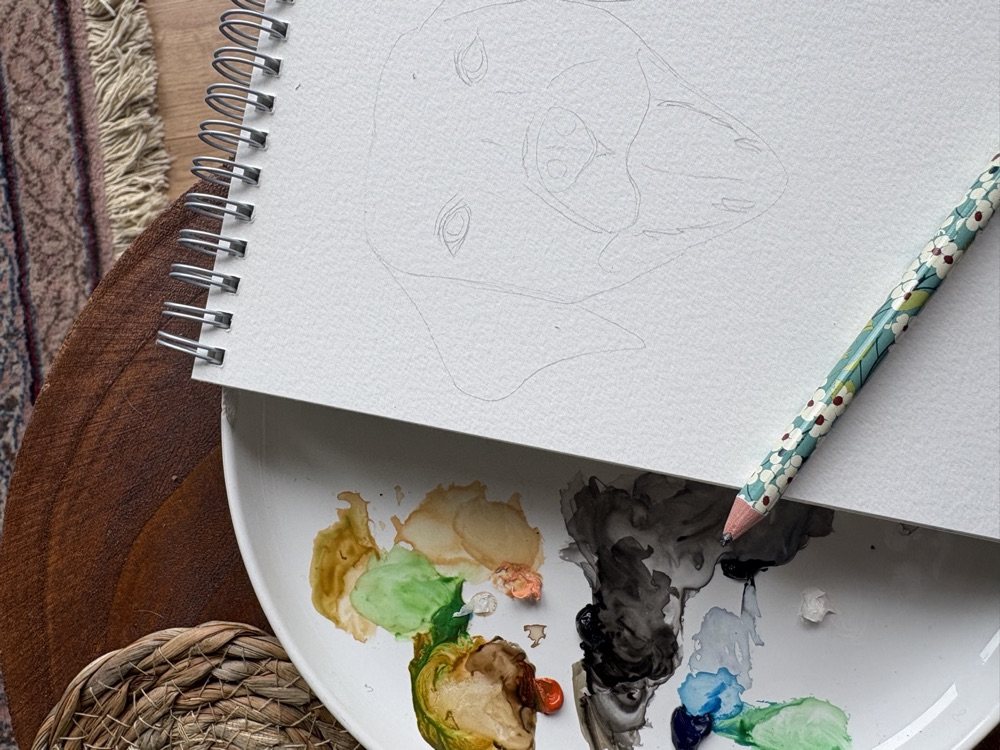Pencil sketch of a yellow Labrador's face on a watercolour pad, paint palette alongside
